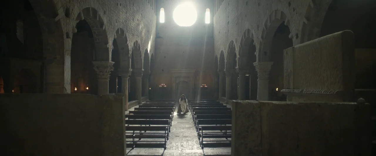 Rosaline (2022) movie still: wide — A stone cathedral nave features rows of dark wooden pews and arched ceiling supports. A woman in a white dress…