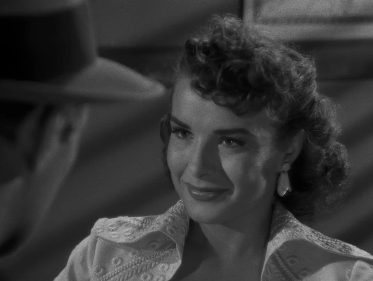 Pickup on South Street (1953) movie still: closeup — A dimly lit interior space features a woman in the foreground and a man. The man wears a fedora…