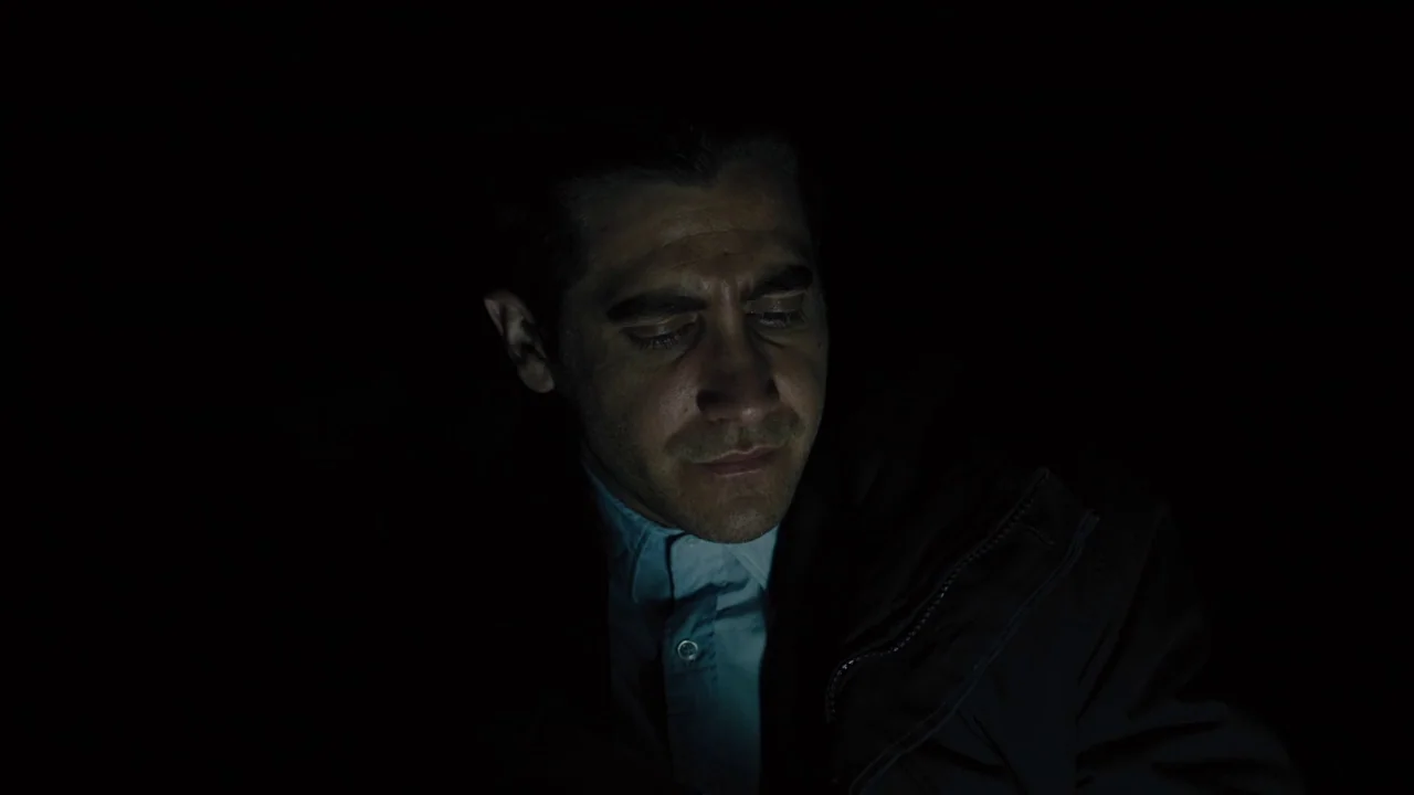 Prisoners (2013) movie still: closeup — A man stands in an environment defined by deep, absolute darkness. He looks down at a light source that…