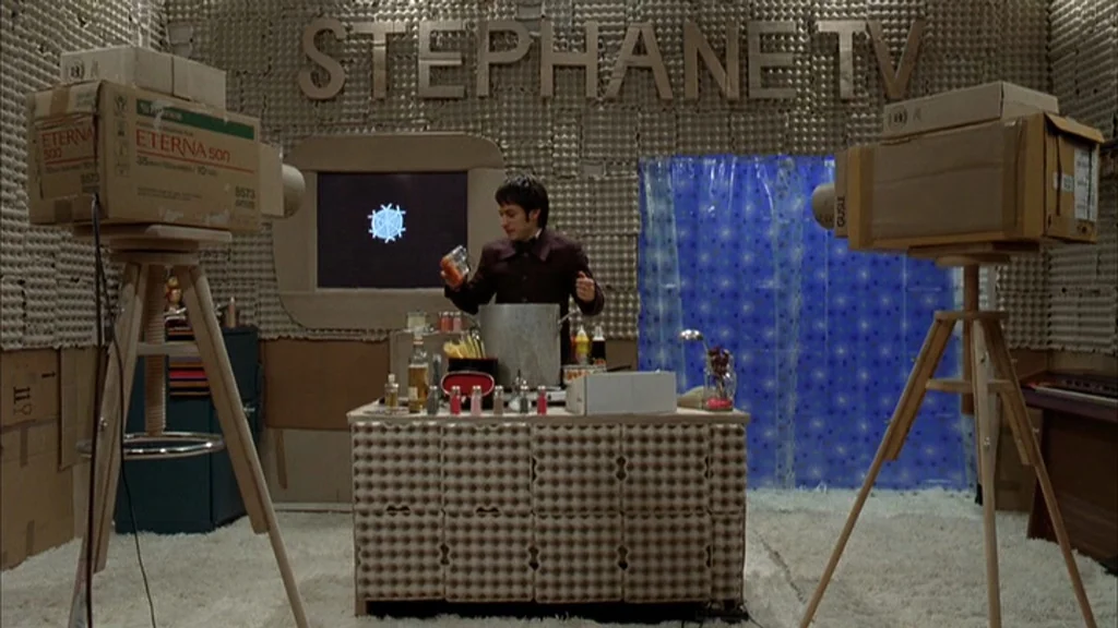 The Science Of Sleep (2006) movie still: wide — A homemade television set constructed from cardboard and egg cartons with white carpet floors. A man…