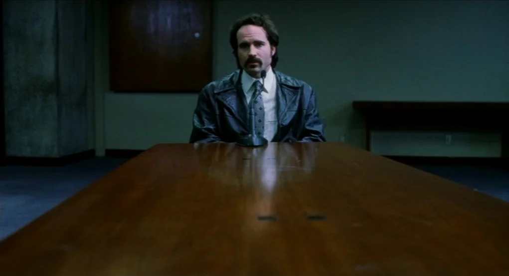 Narc (2002) movie still: medium — A dimly lit room contains a large wooden table and a plain wall behind. A man wearing a leather jacket sits facing…