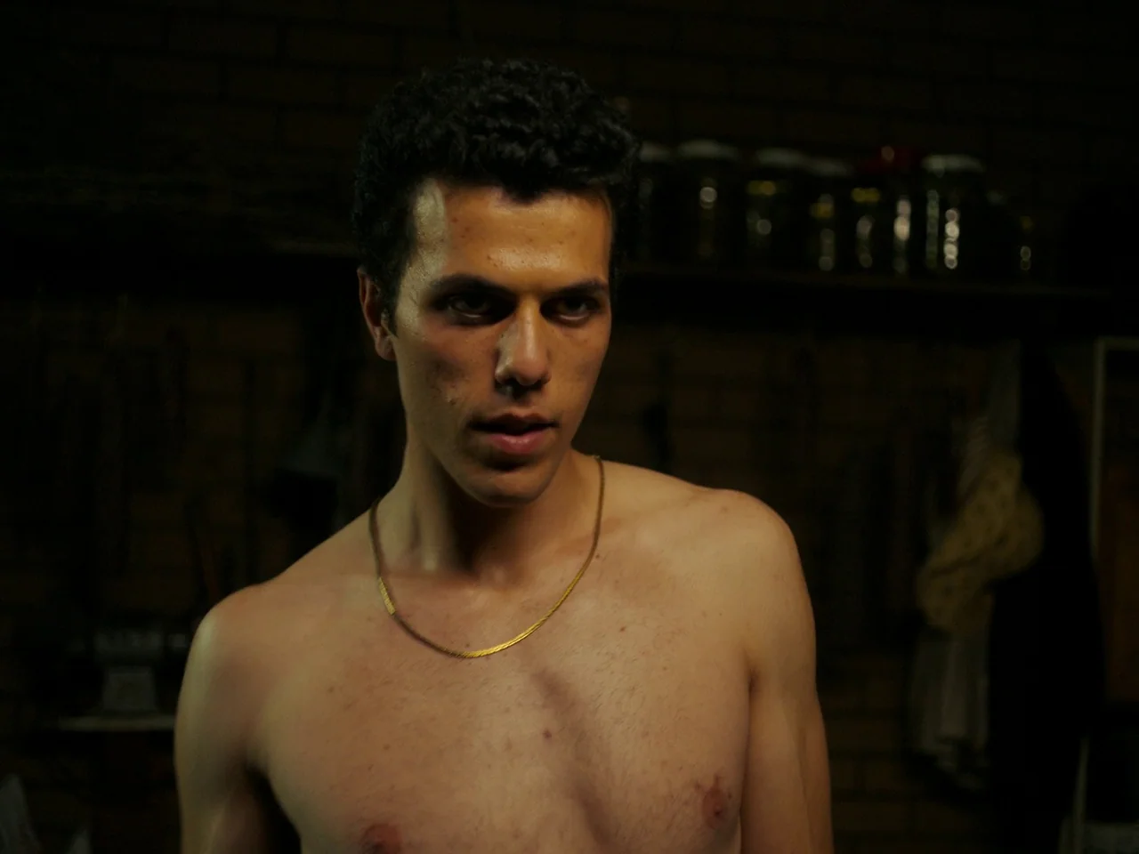 Of An Age (2022) movie still: medium — A dimly lit room with brick walls features rows of glass jars on a shelf. A shirtless young man with curly…