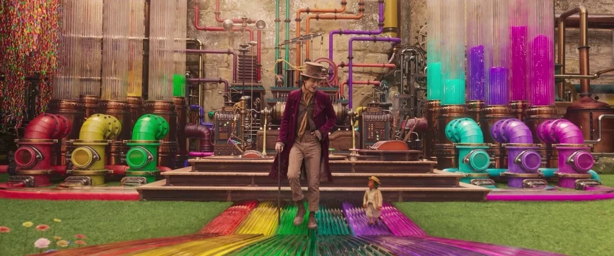 Wonka (2023) movie still: fullbody — A vibrant industrial space filled with multicolored tubes, bubbling vats, and pipes. A tall man and a small man…