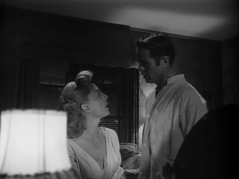 The Killing (1956) movie still: medium — A dark, shadows-filled room features a woman and man engaged in a serious conversation. The woman looks up…