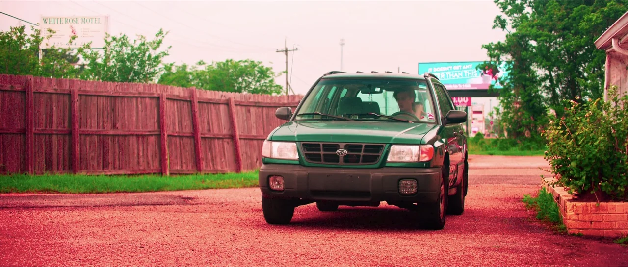 68 Kill (2017) movie still: medium — A green SUV sits on a gravel surface in front of a wooden fence. A young man sits in the driver seat looking…