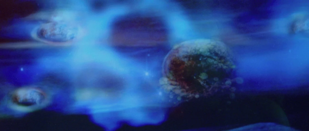 After Blue (2021) movie still: wide — An expanse of dark space filled with blue nebular gas and drifting spherical objects. Small white points of…