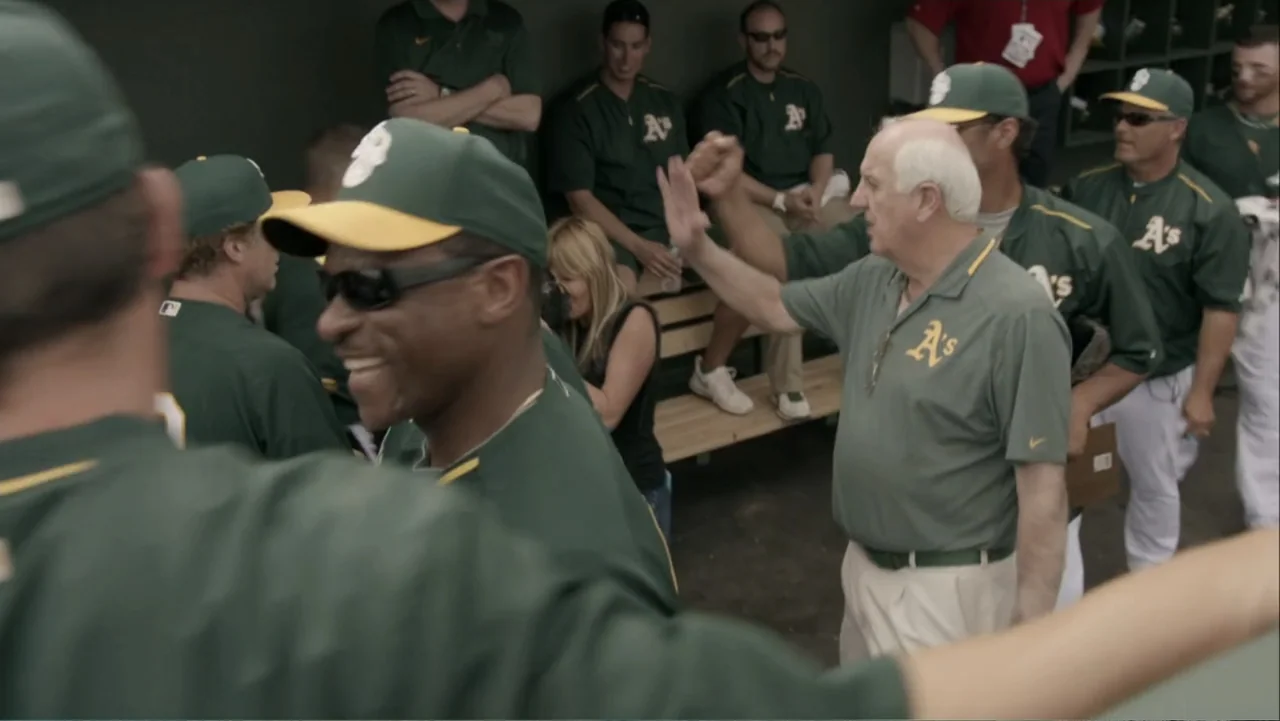 Ferrell Takes the Field (2015) movie still: medium — A professional baseball team dugout with several players seated on a wooden bench. An elderly…