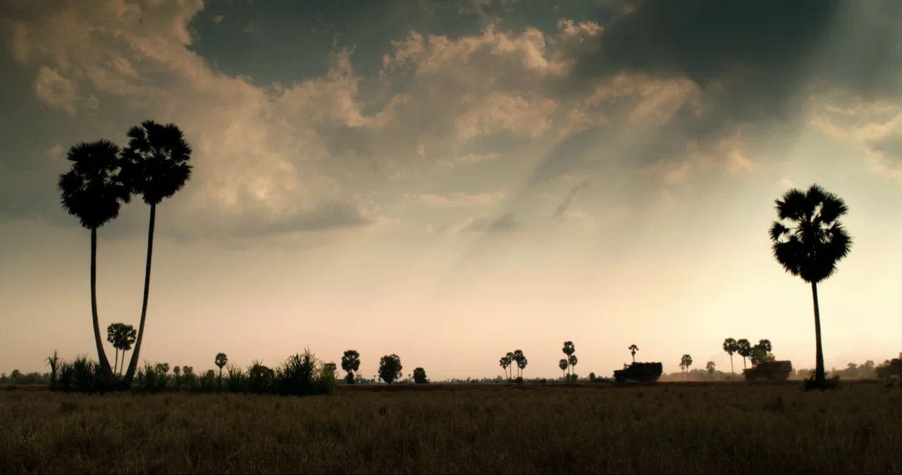 First They Killed My Father (2017) movie still: wide — A vast, flat landscape dotted with tall palm trees under a heavy, overcast sky. Two military…
