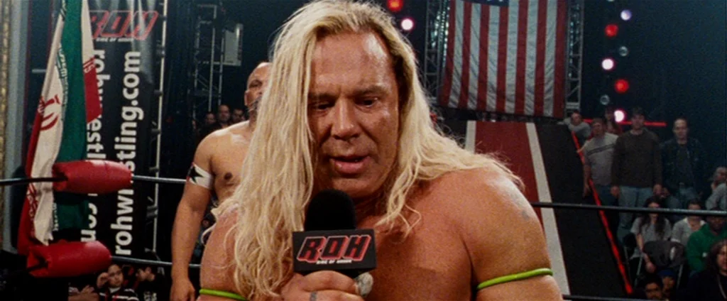 The Wrestler (2008) movie still: medium — A brightly lit indoor arena features a wrestling ring decorated with flags and banners. A blonde wrestler…