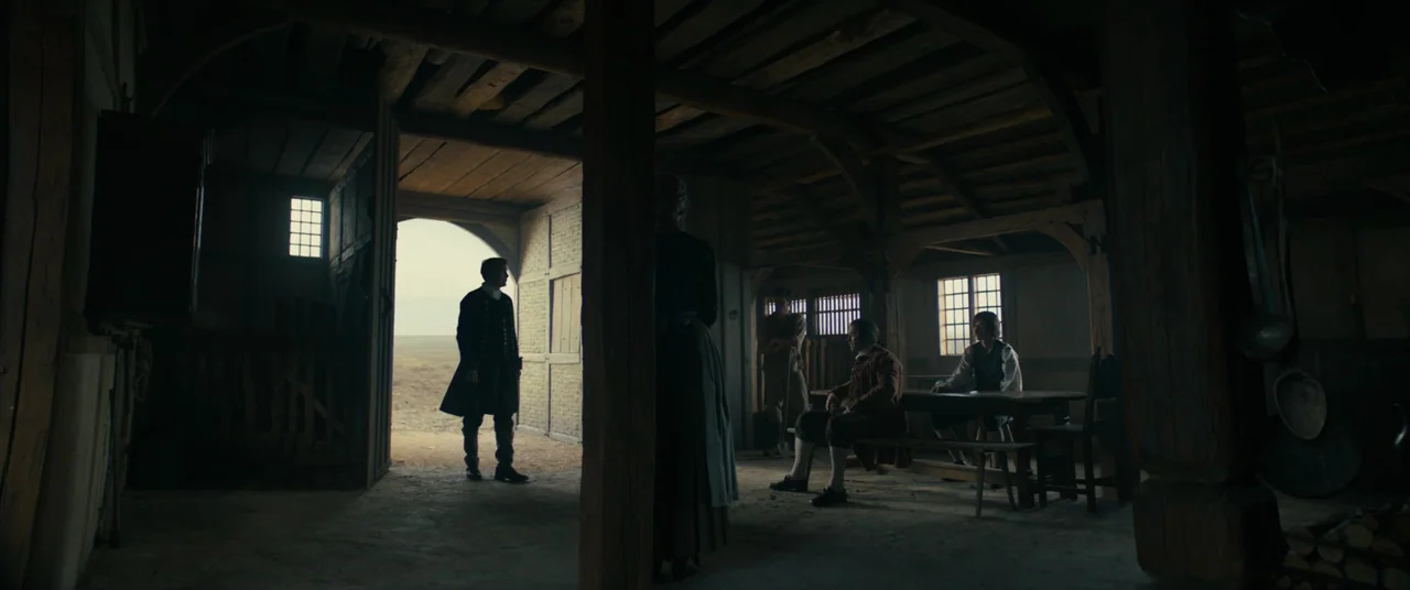 The Promised Land (2023) movie still: wide — A dark timber-framed structure serves as a meeting place with a large open doorway. Several individuals…