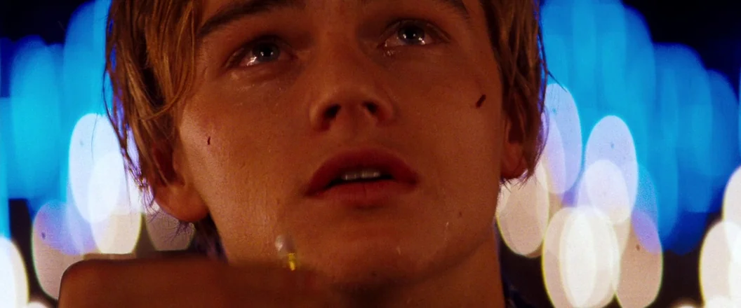 Romeo + Juliet (1996) movie still: closeup — A close-up shot focuses on a young man standing against a blurred nighttime backdrop. The background…