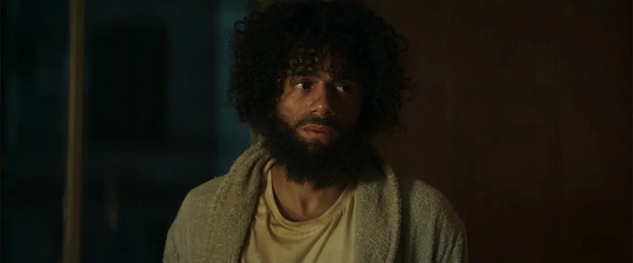 McWalter (2025) movie still: medium — A dimly lit room with plain walls creates a dark and minimalist indoor environment. A bearded man with curly…