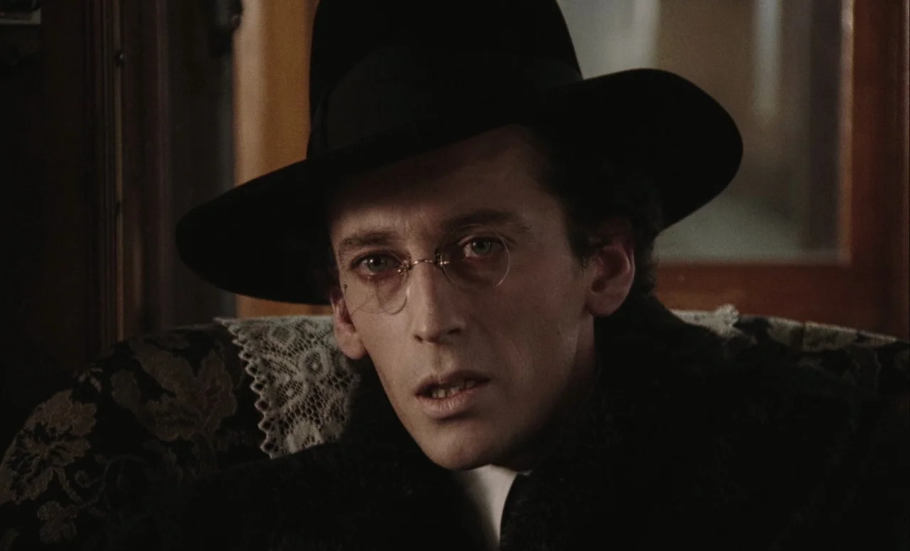 Mahler (1974) movie still: closeup — A close view of a man seated in an ornate, dark patterned upholstery seat. He wears a formal black hat and…