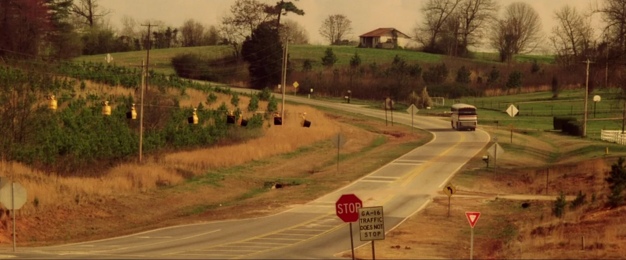 Kiss Kiss Bang Bang (2005) movie still: wide — A rural paved road winds through grassy fields and sparse trees under overcast skies. A passenger bus…