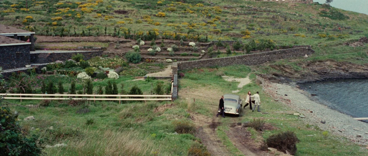 Death Walks On High Heels (1971) movie still: wide — A coastal landscape features a stone house with terraced hills and a rocky beach. Three people…