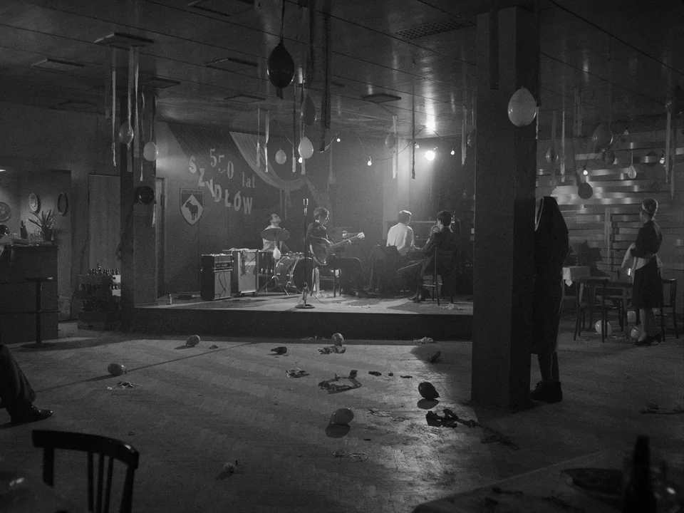 Ida (2013) movie still: wide — A spacious dance hall features a low wooden platform with a small musical band. Scattered balloons hang from the…