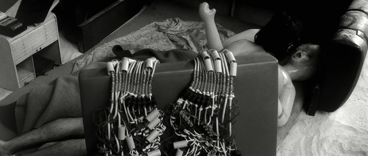 The Last Battle (1983) movie still: medium — A dark, cluttered indoor space contains mannequin limbs and torso fragments on a bed. Electronic…