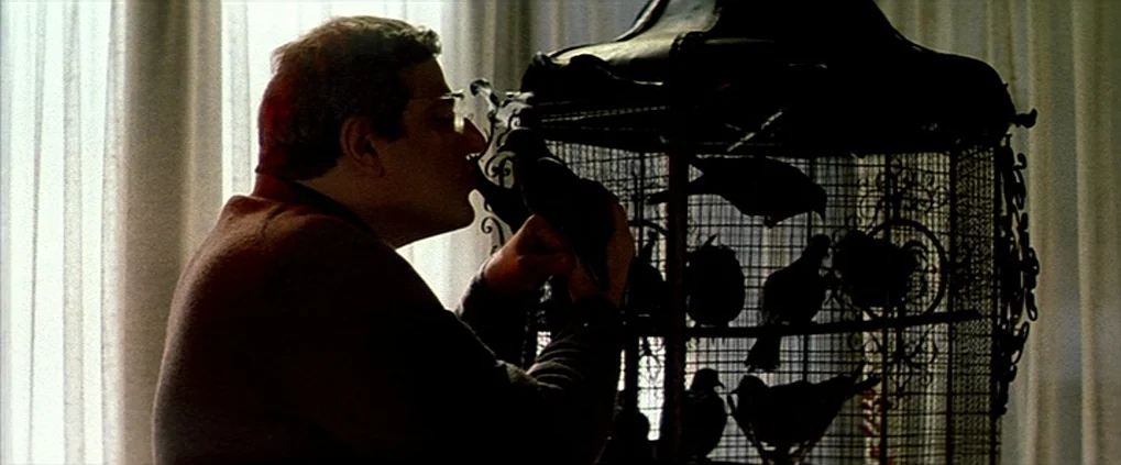 Anguish (1987) movie still: medium — A man stands before a large, ornate wrought iron birdcage filled with several pigeons. He holds one bird close…