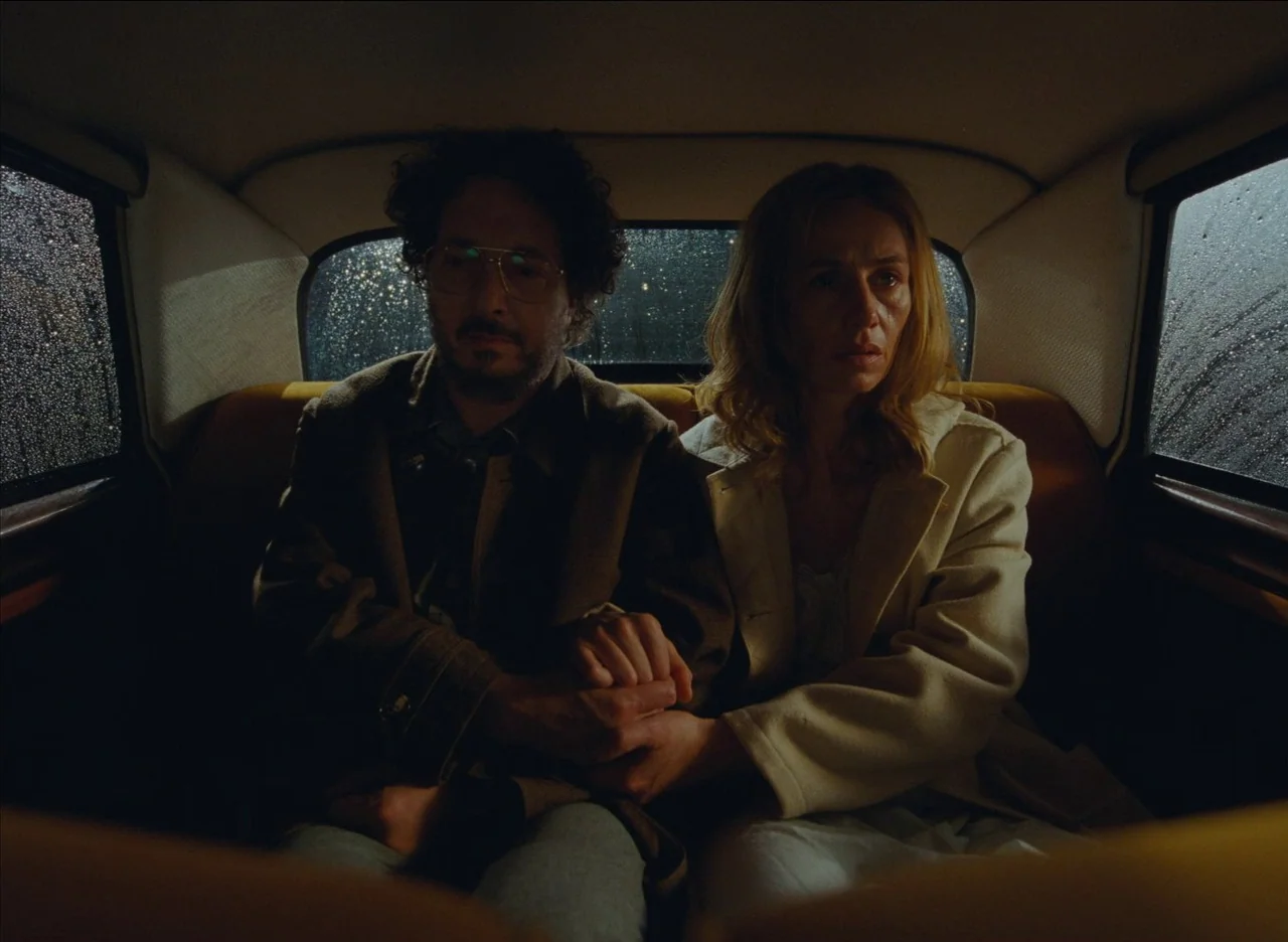 The French Dispatch (2021) movie still: medium — A dark car interior at night with rain streaks on the windows. A couple sits together in the…