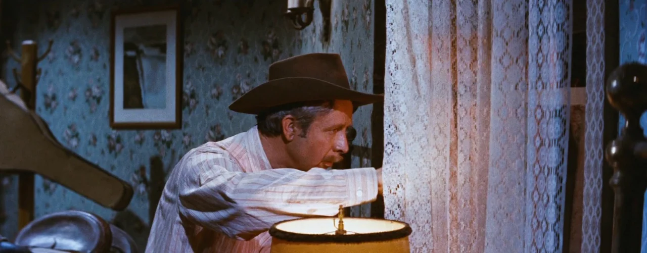 Bus Stop (1956) movie still: medium — A dark room decorated with patterned vintage floral wallpaper features a wooden bed frame. A man wearing a…