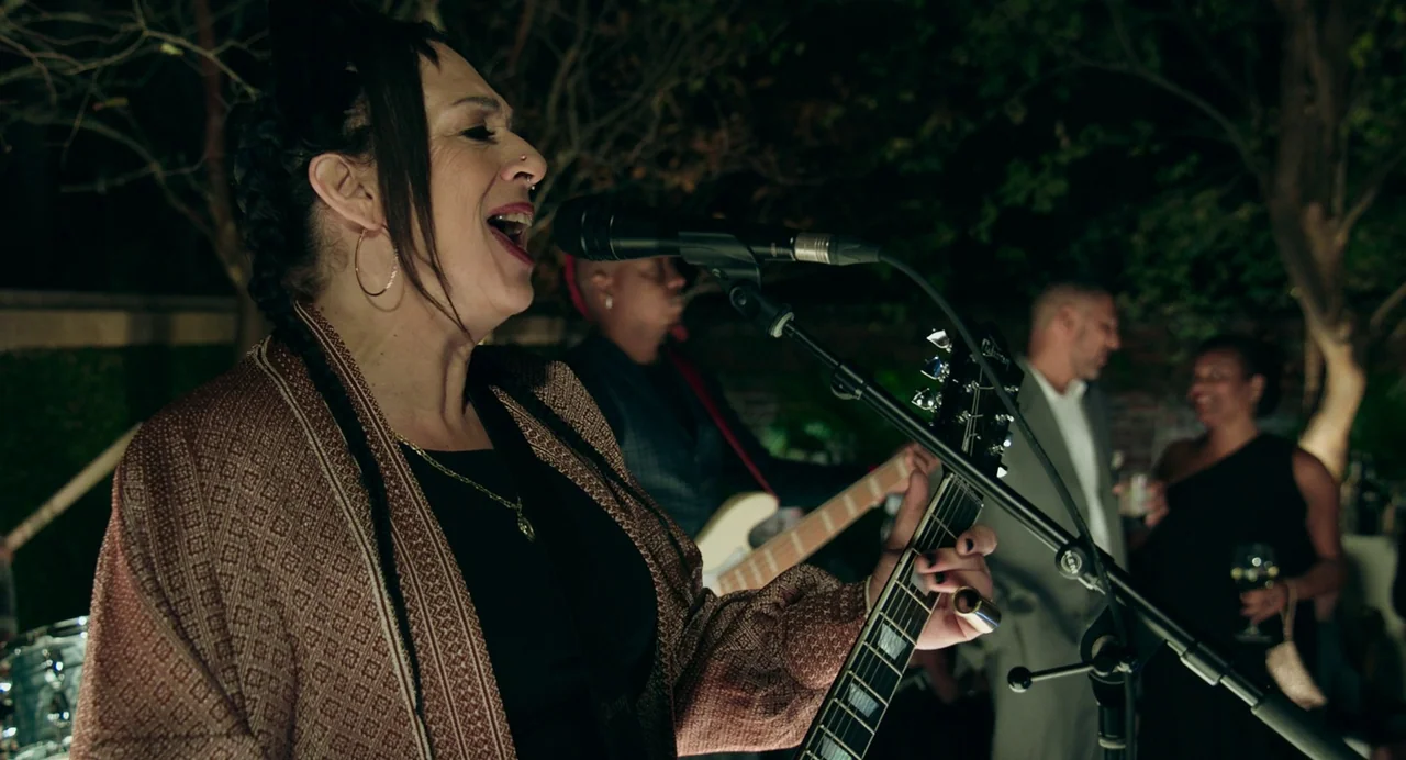 Deep Water (2022) movie still: medium — An outdoor evening event features a live band performing under dark trees. A female singer performs at the…