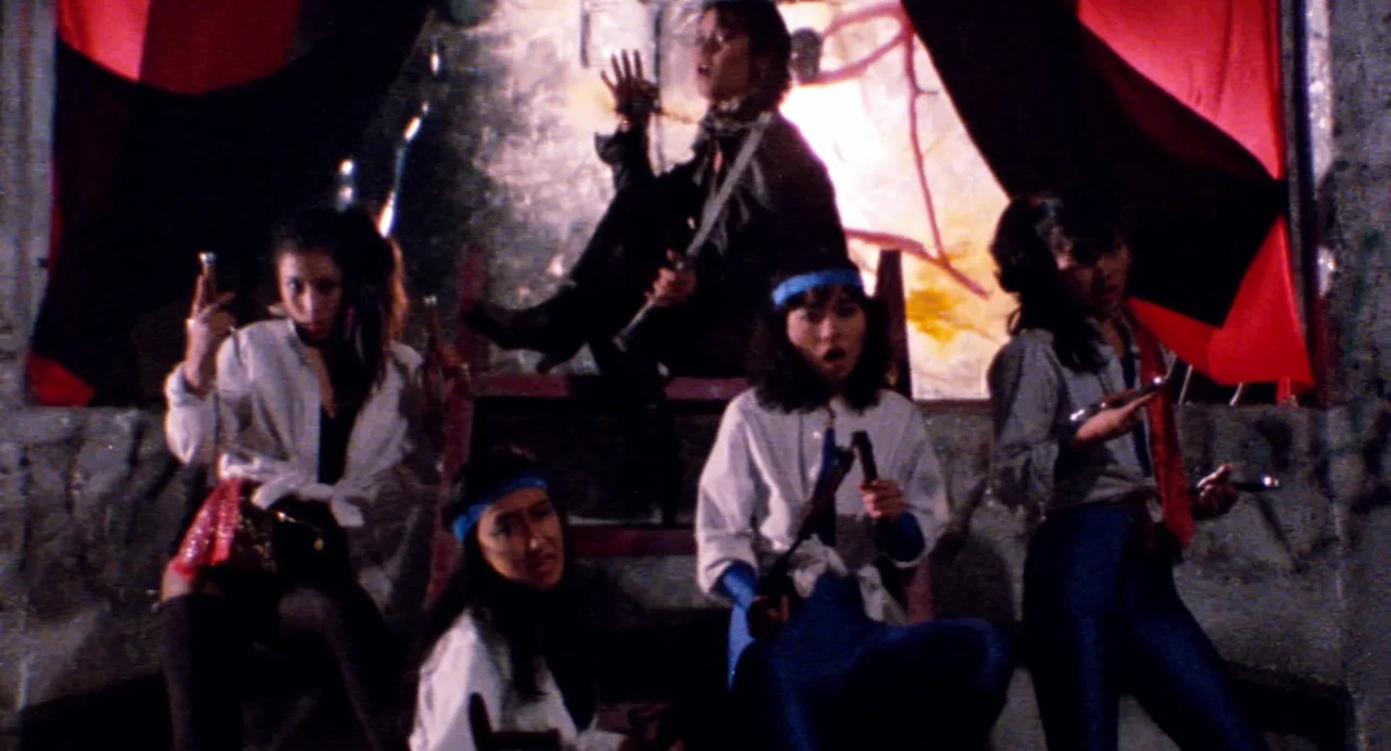 Burst City (1982) movie still: fullbody — A dimly lit room with raw concrete walls and draped red fabric partitions. Five women pose holding various…