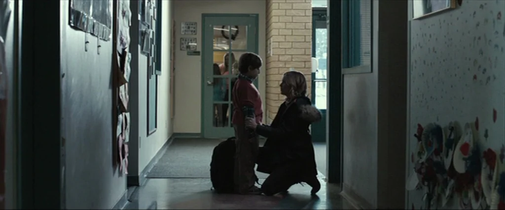 Clown (2014) movie still: fullbody — A school hallway features brick walls covered with colorful children's art projects and displays. A woman…
