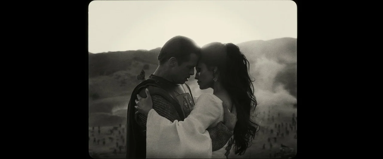 Babylon (2022) movie still: medium — A man in medieval armor and a woman in a gown embrace on a hill. In the distance, smoke rises from an expansive…