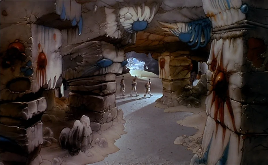 Life Is A Bed of Roses (1983) movie still: wide — A subterranean chamber features thick stone pillars adorned with blue growths and red-brown…
