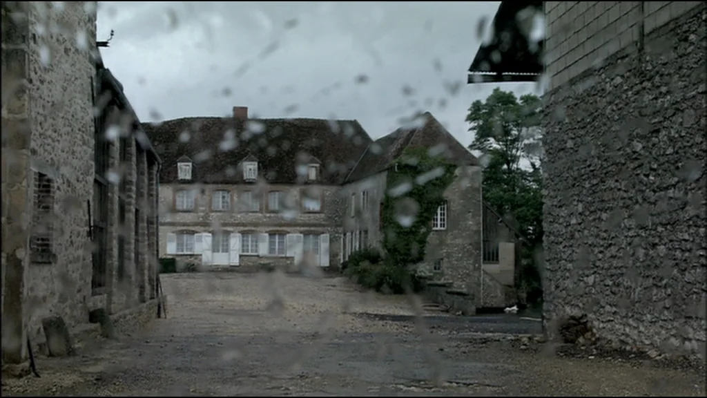 Caché (Hidden) (2005) movie still: establishing — A stone-walled farm courtyard features a large, rustic country house with multiple windows.…