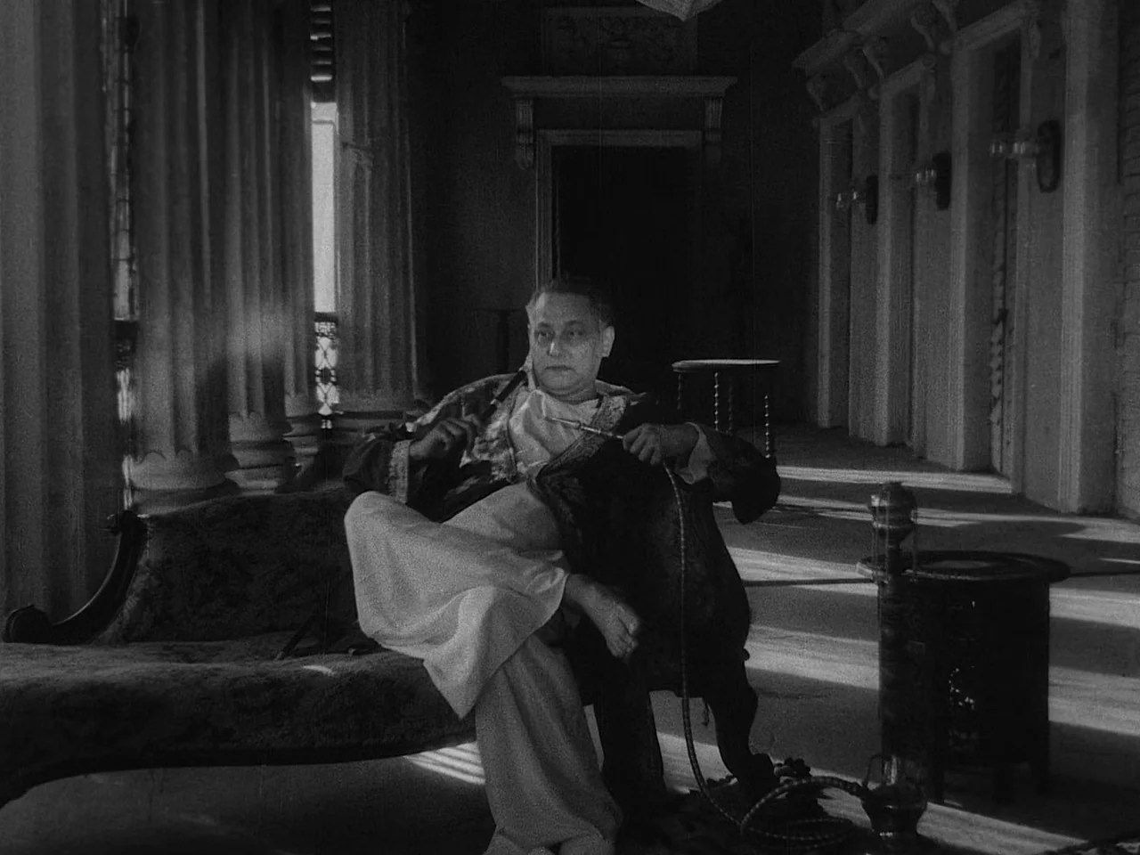 The Music Room (1958) movie still: medium — A spacious veranda with large columns features tiled floors and dark wooden doorways. A man in…