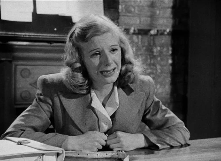 The Blue Lamp (1950) movie still: medium — An interior space features a brick wall backdrop and a smooth desk surface. A woman in a structured suit…