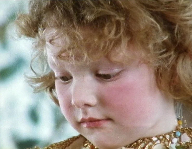 The Tempest (1979) movie still: closeup — A close-up shot focuses on a child with curly hair and a decorative necklace. The child looks down while…