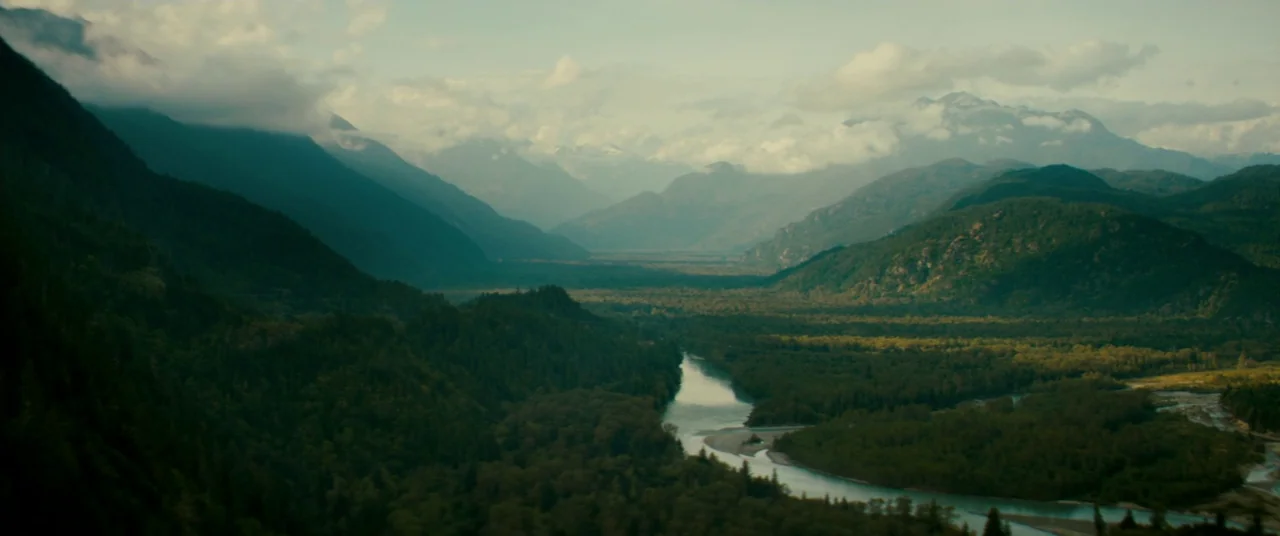 Horns (2013) movie still: establishing — A vast river valley stretches between dense, tree-covered mountain ranges under a cloudy sky. A winding…