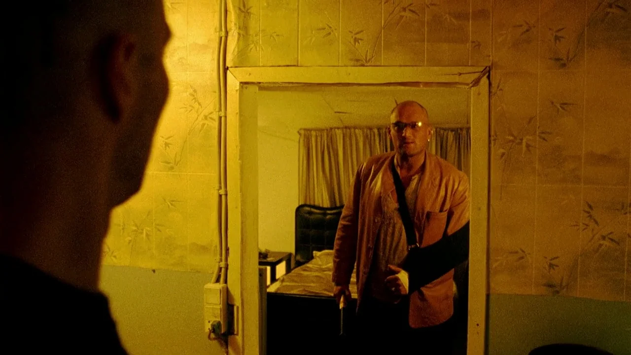 Pusher II (2004) movie still: medium — An interior shot shows a doorway leading to a dim, cluttered bedroom space. A bald man wearing a sling stands…