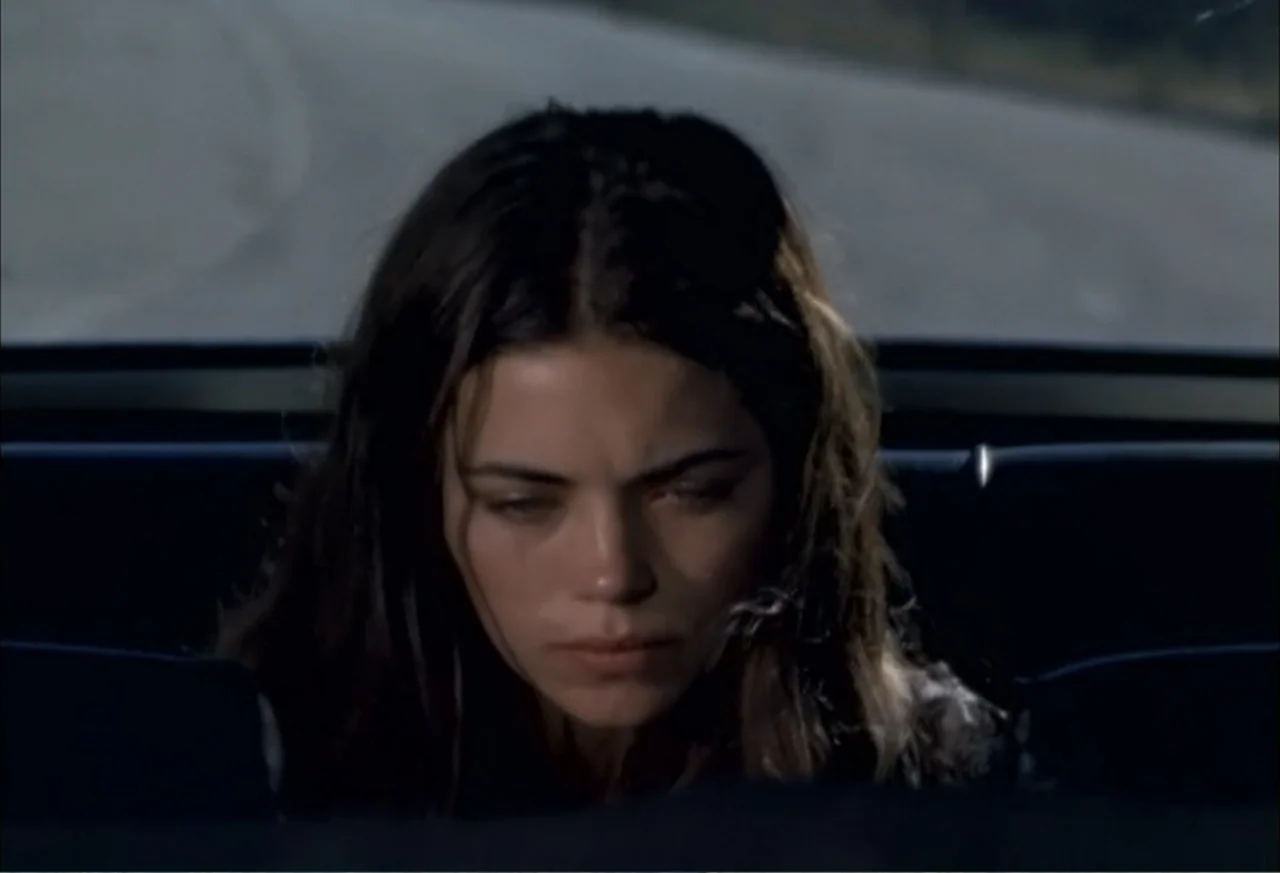 Black Cat Run (1998) movie still: closeup — A woman sits in the passenger seat of a vehicle moving along a road. The woman stares downward while the…
