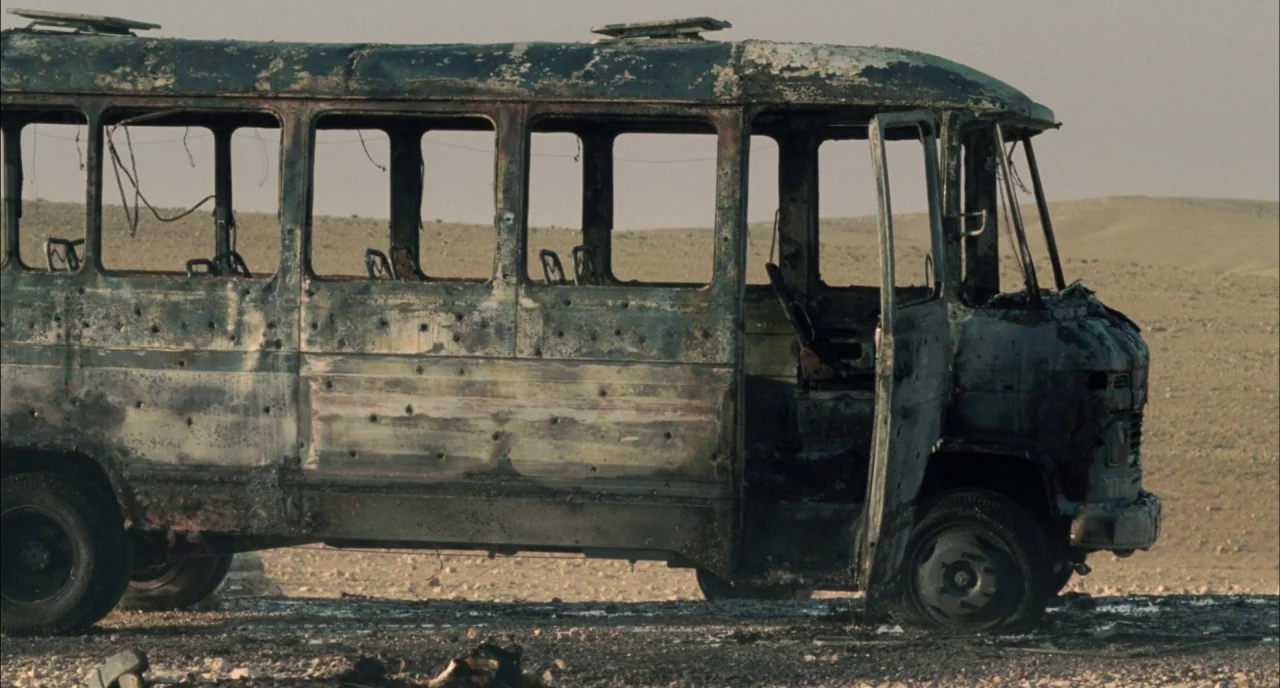 Incendies movie still: wide — A scorched and bullet-riddled bus sits abandoned in a barren, dusty desert landscape. The vehicle is missing glass and…