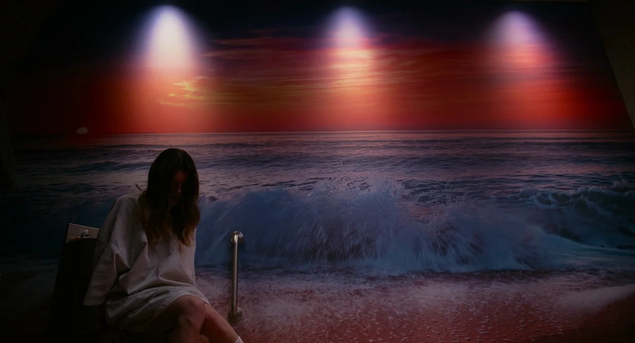 Fresh (2022) movie still: medium — A bathroom wall is covered with a large mural of a sunset ocean scene. A woman sits on the toilet in front of the…