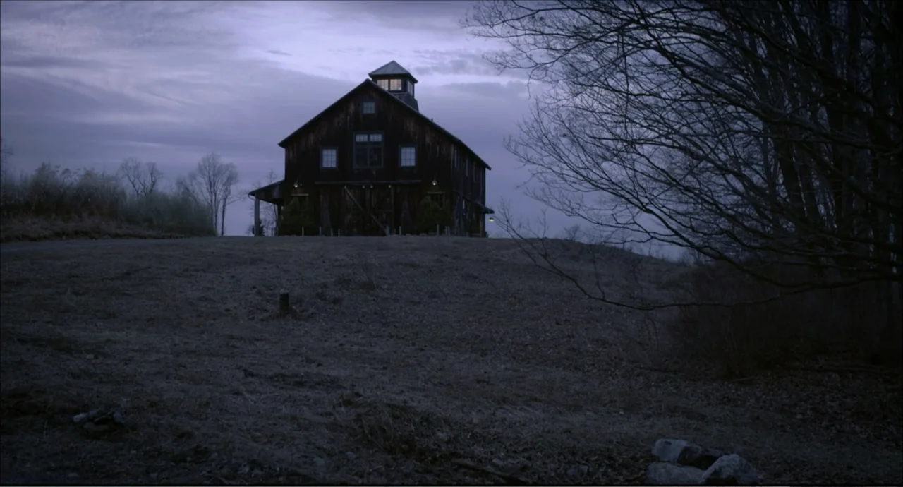 Freedom (2024) movie still: establishing — A dark wooden barn stands atop a grassy hill during the blue hour. Bare tree branches frame the right…