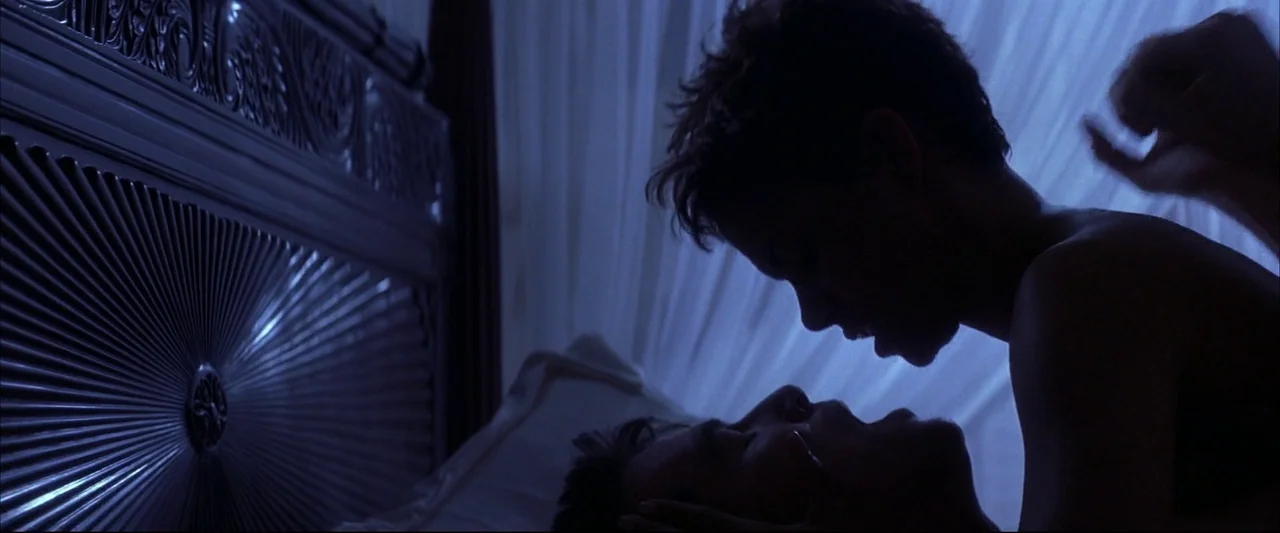 Die Another Day (2002) movie still: medium — A dark bedroom features a large, ornate headboard with a metallic circular pattern. A couple lies in…