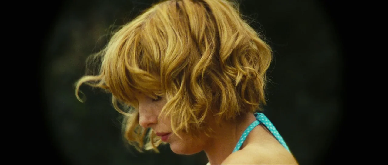 Eden Lake (2008) movie still: closeup — A close up of a blonde woman in a park with dark trees blurred behind. The woman wears a blue polka dot…