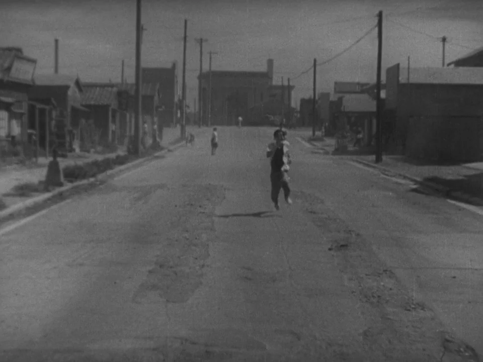 Stray Dog (1949) movie still: wide — A dusty, unpaved road runs between simple wooden structures under an overcast sky. A young child runs directly…
