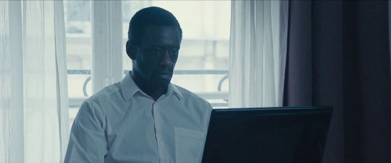 Simon Killer movie still: medium — A man sits before a laptop in a room with light curtains over a window. He focuses his gaze on the glowing screen…