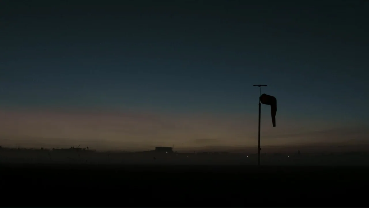 Transfers (2024) movie still: establishing — An open field at dusk features a distant low horizon with small silhouetted structures. A windsock on a…
