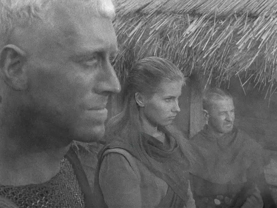 The Seventh Seal (1957) movie still: medium — Three people stand outside near a thatched roof structure in a rural landscape. The man in the…