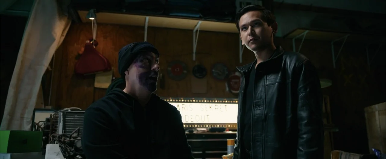 Camp Hideout (2023) movie still: medium — A cluttered garage workspace contains various tools and a glowing marquee sign with text. Two men stand in…