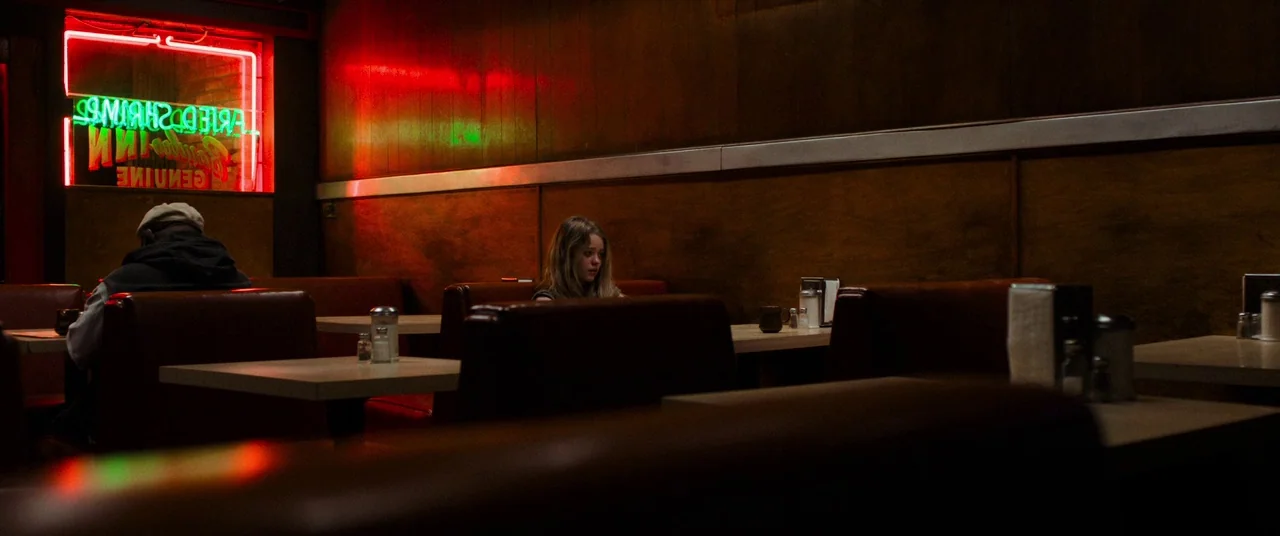 Destroyer (2018) movie still: wide — A dark, wood-paneled booth area of a diner with a bright neon window sign. A woman sits alone in the distance…