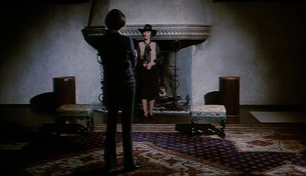 Harold and Maude (1971) movie still: fullbody — A stone room features a large central fireplace and an ornate patterned carpet. Two women stand…