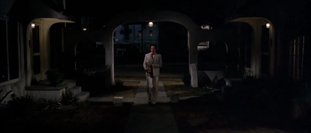 The Fortune (1975) movie still: wide — A night view of an apartment complex courtyard featuring symmetric stucco archways. A man wearing a light…