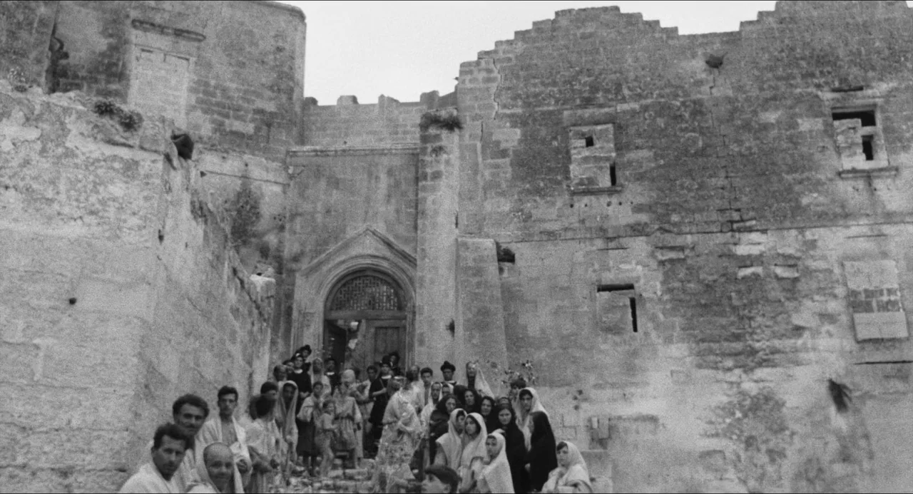 The Gospel According To St. Matthew (1964) movie still: wide — Ancient, textured stone structures surround a weathered arched doorway in a desolate…